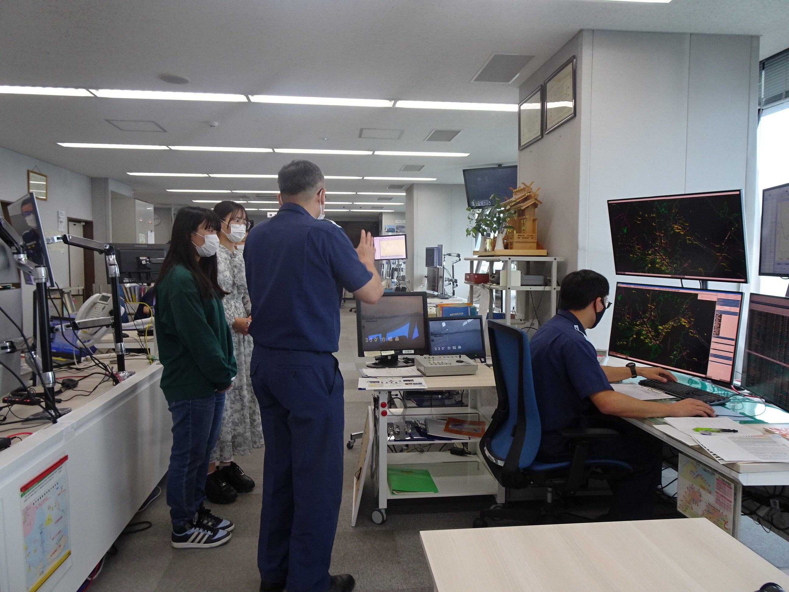 管制官室の管制卓に座って、レーダーやAIS、高性能カメラ等について説明している様子